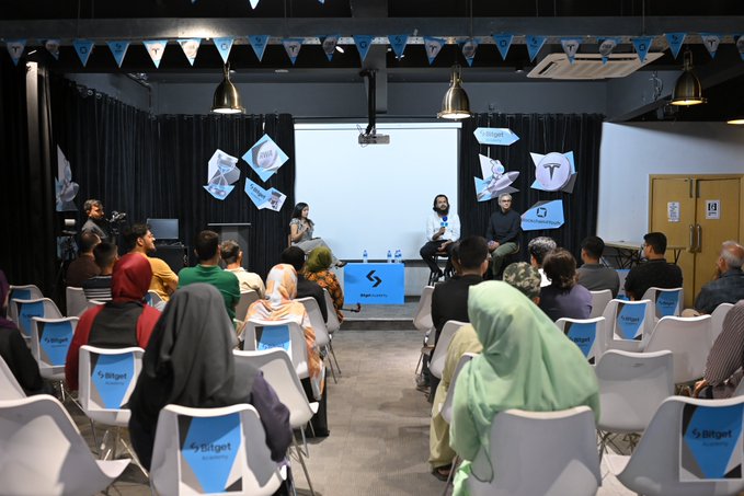 A Bitget Academy event titled "Crypto Experience Month in SA" takes place in a conference room, with two panelists seated on stage in front of a projection screen, while an attentive audience—including women in hijabs—sits in white chairs branded with the Bitget logo, reflecting the post's focus on education and empowerment of future crypto talent.