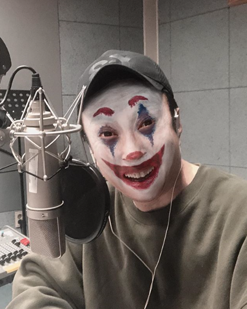 Person wearing a camouflage cap and a Joker-style clown makeup (white face with blue and red accents), smiling in front of a studio microphone.