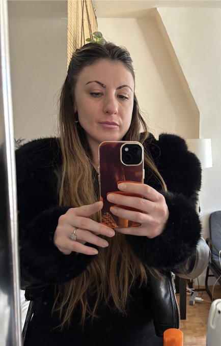 A woman takes a mirror selfie wearing a black fuzzy jacket, holding a phone with a tortoiseshell case in a bright room.