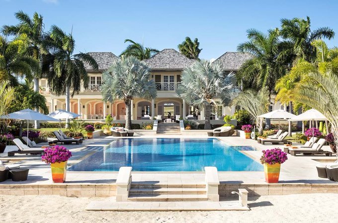 A luxurious tropical resort featuring a large swimming pool, palm trees, lounge chairs, and a grand villa-style building under a clear blue sky.