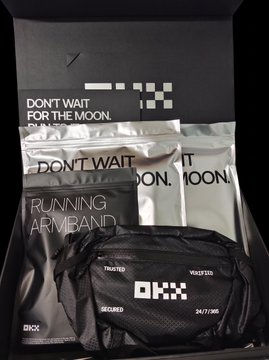 A black OKX branded box and running armband with the text 'DON'T WAIT FOR THE MOON' and 'RUNNING ARMBAND SECURED 24/7/365'.