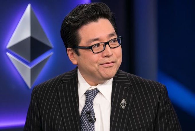 A man in a pinstripe suit, tie, and glasses smiles in front of an Ethereum logo backdrop, wearing an Ethereum lapel pin.