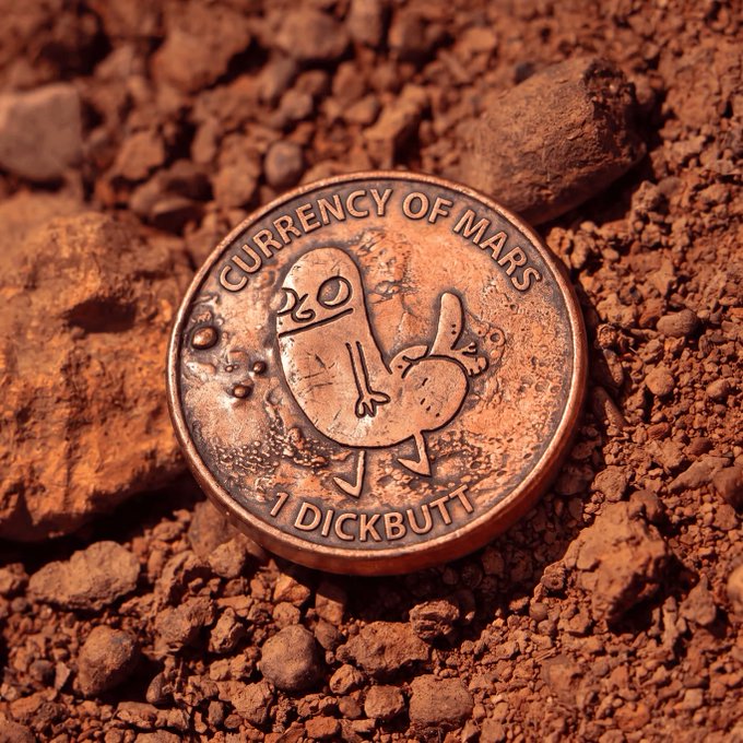 A copper-colored novelty coin labeled 'CURRENCY OF MARS' and '1 DICKBUTT' resting on reddish rocky soil.