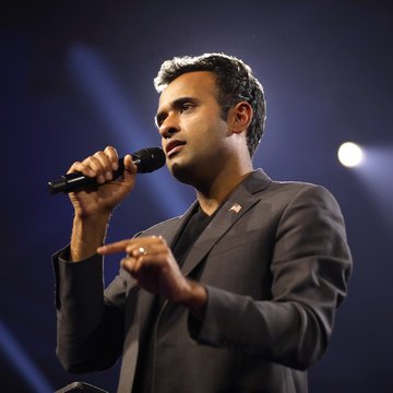 The image features a man wearing a suit and tie standing on stage with his hand raised into the air while holding a microphone. The background of the image shows a dark blue color, which contrasts with the man's attire. There are also some lights in the background that illuminate the scene. The post about Vivek Ramaswamy is written at the bottom right corner of the image.