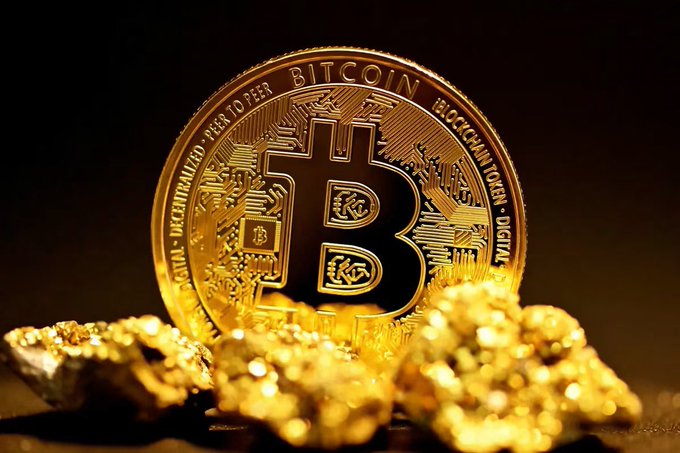 A close-up, warm-lit photograph of a physical gold Bitcoin coin standing upright on a bed of gold nuggets, with the inscription 'BITCOIN DIGITAL DECENTRALIZED PEER TO PEER BLOCKCHAIN TOKEN' visible around the edge.