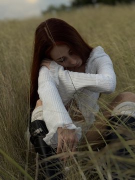 A young woman with long auburn hair, wearing a cream knit sweater, rests her head on her arms while sitting in a golden field of tall grass at golden hour, gazing warmly at the camera. The serene, contemplative mood of the image echoes the post's reflection on the elusive and precious nature of happiness.