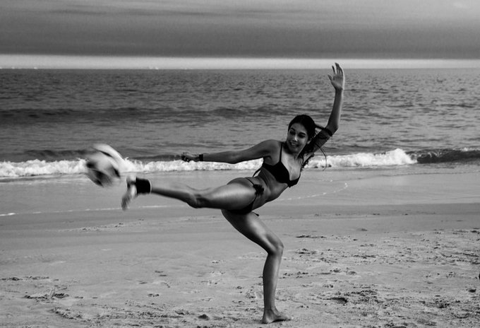 A black and white photograph captures a woman in a bikini on a sandy beach, mid-motion as she appears to be kicking a soccer ball while balancing on one leg, with ocean waves in the background.