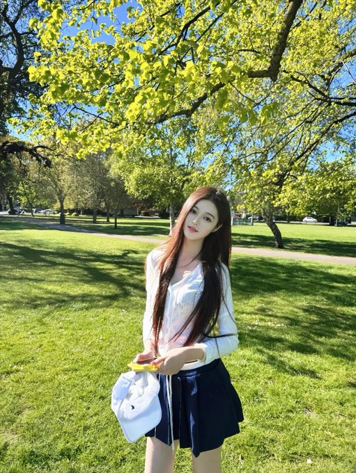A young woman with long dark hair, wearing a white cardigan and navy skirt, stands in a sunlit park holding a leaf, perfectly capturing the fresh essence of a cheerful "Good Morning." The lush green trees and bright sunlight overhead enhance the warm, uplifting greeting.