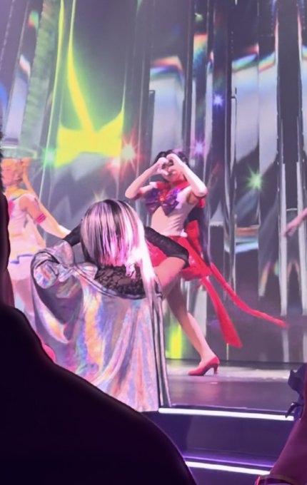 A performer on stage strikes a pose with hands forming a heart shape above her head, wearing a red outfit and long red ribbons, with colorful stage lighting and a star-shaped backdrop visible behind her.