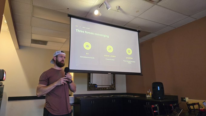 A bearded man in a maroon shirt and white cap speaks into a microphone while presenting a slide titled 'Three forces converging' listing AI, State Power, and Crypto.