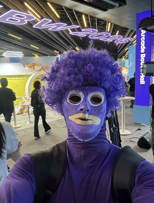 A person dressed in a purple bodysuit with a curly purple wig and a painted face mask takes a selfie at what appears to be a 'BM 2026' (Bilibili Macro Link) event, with a Hamtaro mascot display visible in the background.