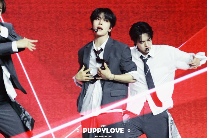 The image features two young men dressed in suits, one wearing a tie and the other wearing sunglasses. They are standing next to each other against a red background with white text that reads "with puppylove" and "allday". The focus of the image is on these two individuals as they pose for the photo.