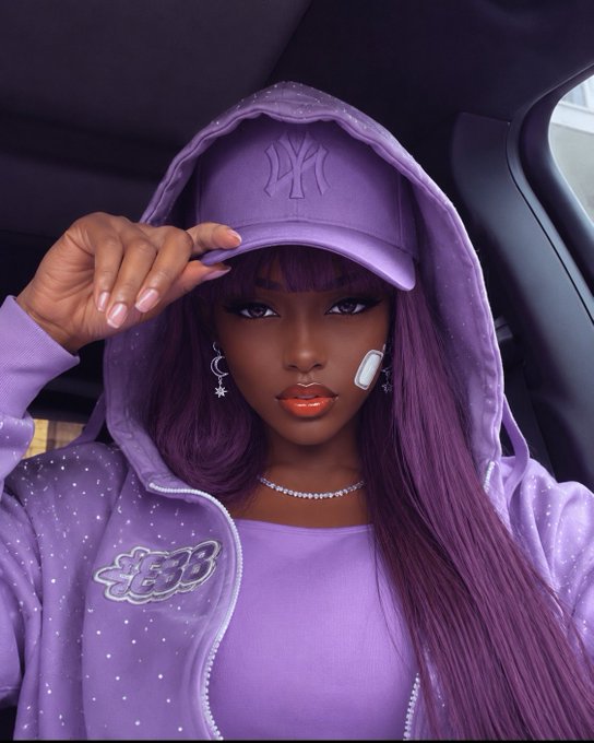 A woman with long purple hair wearing a purple NY Yankees cap, a sparkly purple zip-up hoodie, and a matching top, taking a selfie in a car.