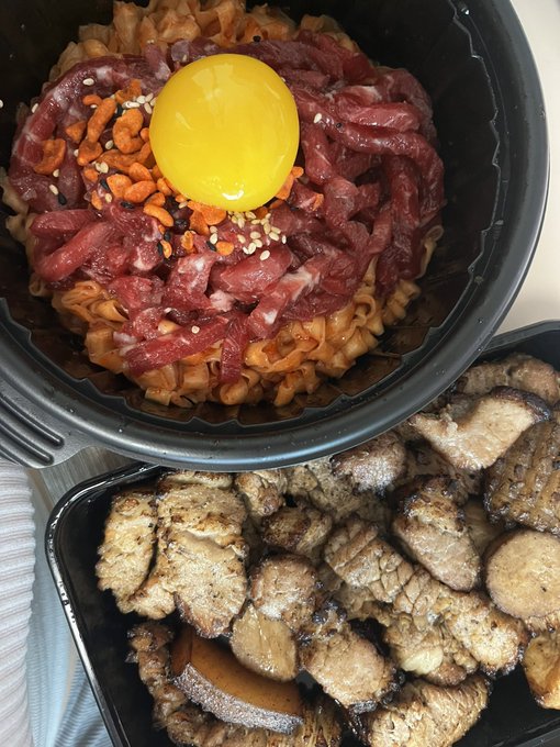 A black bowl containing ramen noodles topped with raw beef, a raw egg yolk, and seasoning, accompanied by a side container of grilled meat.
