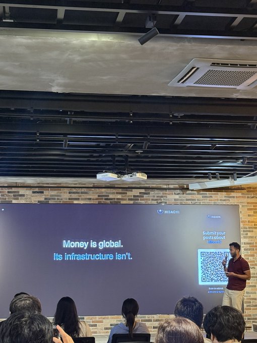 A man in a maroon shirt giving a presentation in front of a large screen displaying the text 'Money is global. Its infrastructure isn't.' alongside a QR code and 'Submit your posts about Bitcoin' text, branded with Hidashi logos.