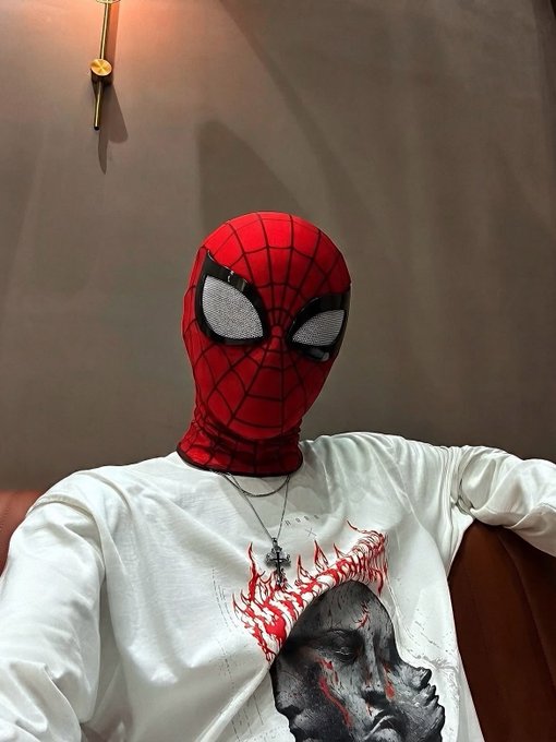 A person wearing a Spider-Man mask and a white long-sleeve shirt with a graphic print, sitting against a brown background.