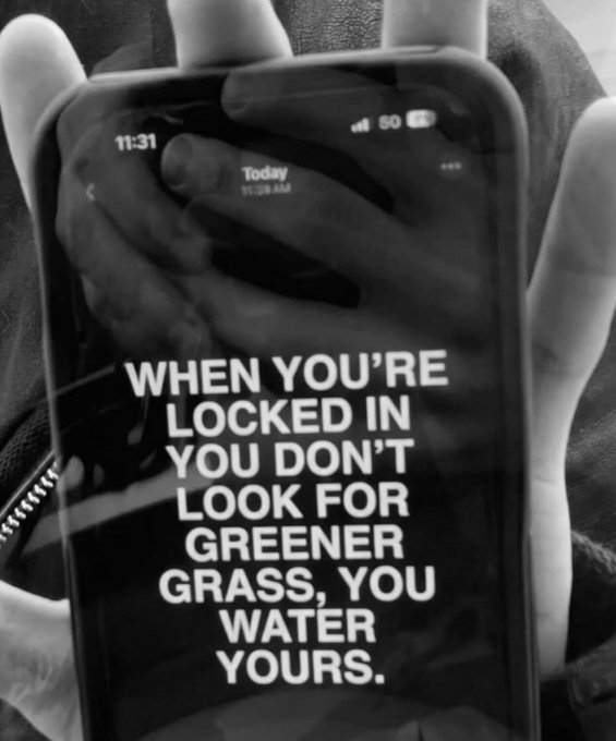 A black and white photo of a hand holding a phone displaying the motivational text: "WHEN YOU'RE LOCKED IN YOU DON'T LOOK FOR GREENER GRASS, YOU WATER YOURS." with the time 11:31 AM shown.