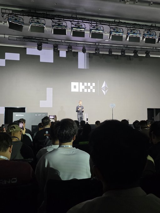 An audience watches a presentation at what appears to be an OKX (OKX logo and Ethereum symbol visible) tech event, with a speaker on stage.