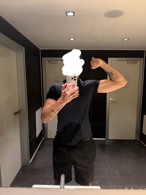 A mirror selfie of a muscular person with tattoos wearing a black t-shirt and shorts, flexing their arm while holding up an iPhone in a tiled bathroom.