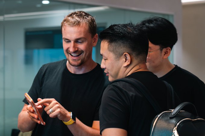 The same scene as the first image - three men in black shirts looking at a smartphone together, with the blonde man smiling while showing something on the screen.