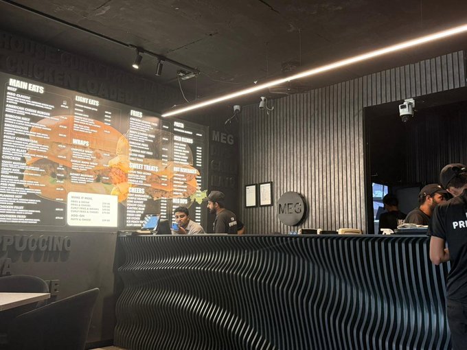 Interior of a fried chicken restaurant showing a menu board with categories like 'MAIN EATS', 'SIDES', and 'SWEET TREATS' alongside staff behind a wavy-textured counter.