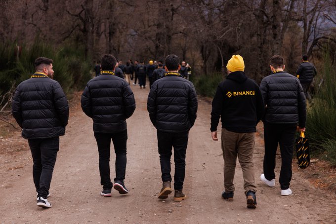 Five individuals walk together down a wooded dirt path, several wearing black jackets displaying the "BINANCE" logo in yellow, embodying the post's theme of community, learning, and growth through their shared forward journey.