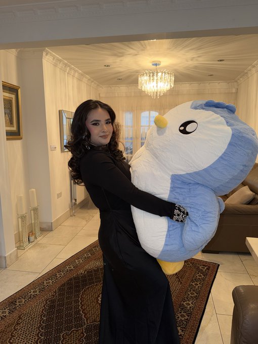 A woman in a black dress stands in an elegant foyer, smiling at the camera while hugging an oversized blue and white Pudgy Penguins plush toy. The image ties to her reintroduction post as a visual showcase of the "lil Pudgy" NFT that she humorously credits with changing her entire life.