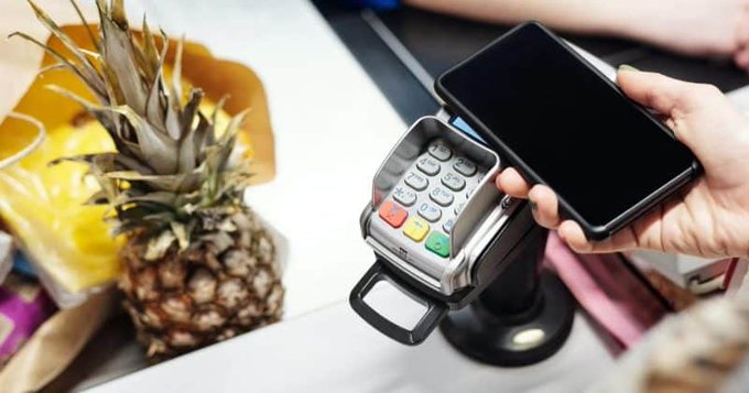 A close-up shows a hand holding a smartphone at a checkout terminal with a PIN keypad, with a pineapple visible at the counter in the background.