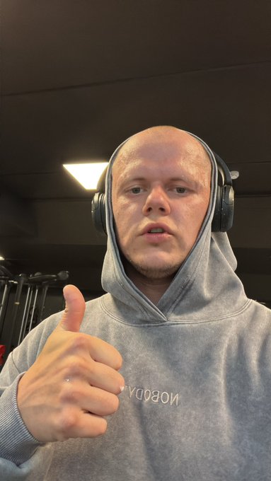 A man wearing a grey 'NOOBDAY' hoodie and large black headphones gives a thumbs-up while looking at the camera.