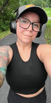 A smiling woman wearing glasses, a black tank top, and headphones takes a selfie outdoors, with a colorful tattoo visible on her shoulder.