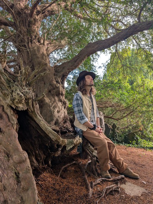 A man wearing a hat, plaid shirt, and tan pants sits on the roots of a large tree in a wooded outdoor setting.