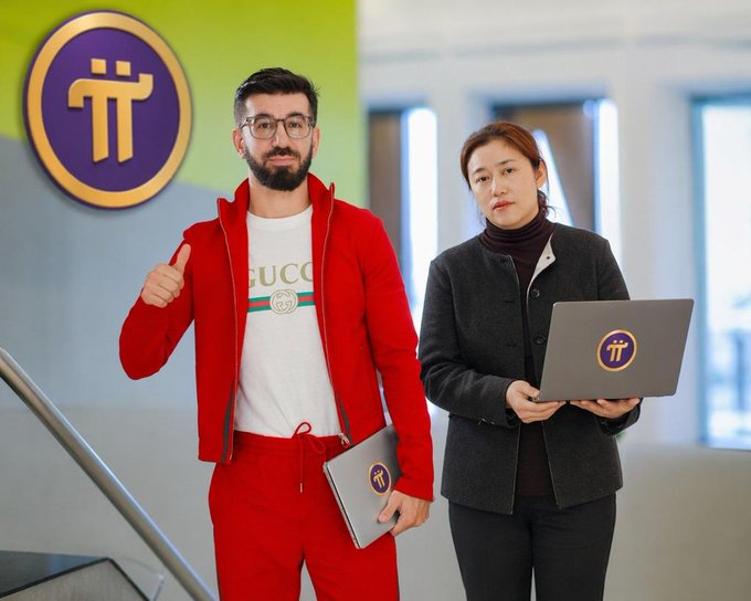 Two people pose in a Pi Network-branded office setting, with a bearded man in a red tracksuit giving a thumbs-up beside a woman in a dark blazer holding a Pi-branded laptop. The image relates to the post by visually representing Pi Network's core team and Pioneers community, which is currently grappling with a v25.2 node update issue causing status resets due to a Stellar Core adaptation conflict.