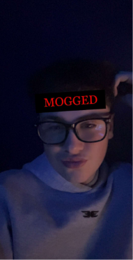 Portrait of a young man with glasses in a dark, blue-lit setting, with the word 'MOGGED' displayed across his forehead in red text.