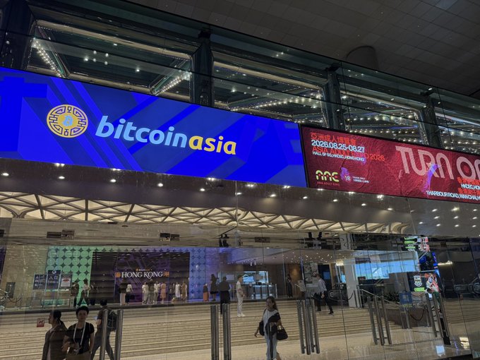 A massive digital billboard at Hong Kong's convention venue advertises "bitcoin asia" alongside the "TURN ON" Asian Adult Expo (scheduled for August 25-27, 2026), humorously confirming the scheduling coincidence between the two major events sharing the same location.