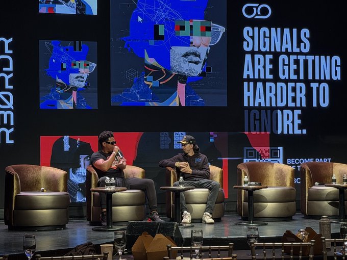 Two men seated in brown leather chairs on a stage at a 'REORDER' conference or event, with a large screen behind them displaying artistic portraits and the tagline 'SIGNALS ARE GETTING HARDER TO IGNORE.'