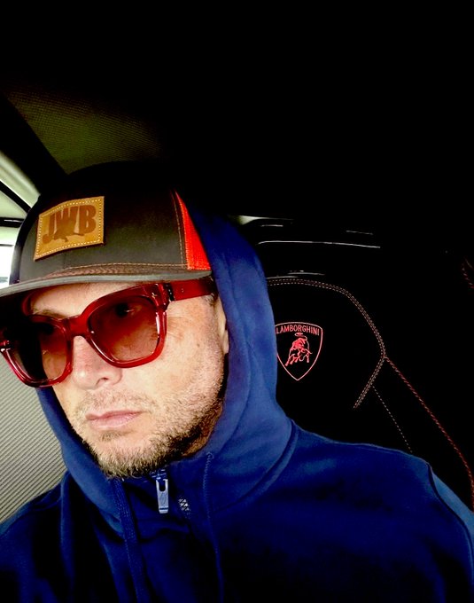 Jason wears a brown leather JWB cap, red sunglasses, and a blue hoodie in the driver's seat of a Lamborghini, perfectly matching his post about spending money on "crazy shit" with the vehicle's logo visible on the seat.