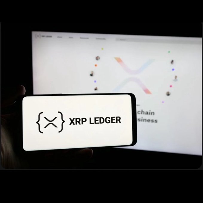 A smartphone displaying the 'XRP LEDGER' logo with its distinctive bracket-X symbol is held up in front of a computer monitor showing the XRP website.
