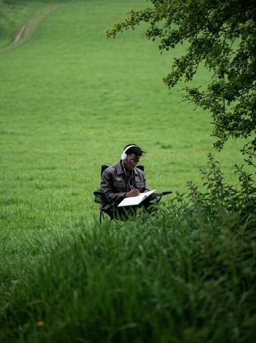 A serene scene of a person wearing headphones sitting in a folding chair in a lush green field, drawing or writing on a pad.
