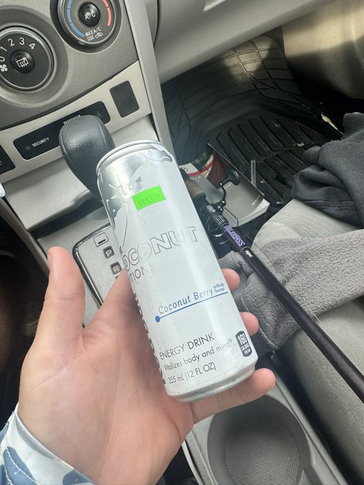 A hand holding a silver can of 'Coconut Nation Coconut Berry Energy Drink' (355 mL) inside a vehicle, with the center console and gear shift visible in the background.