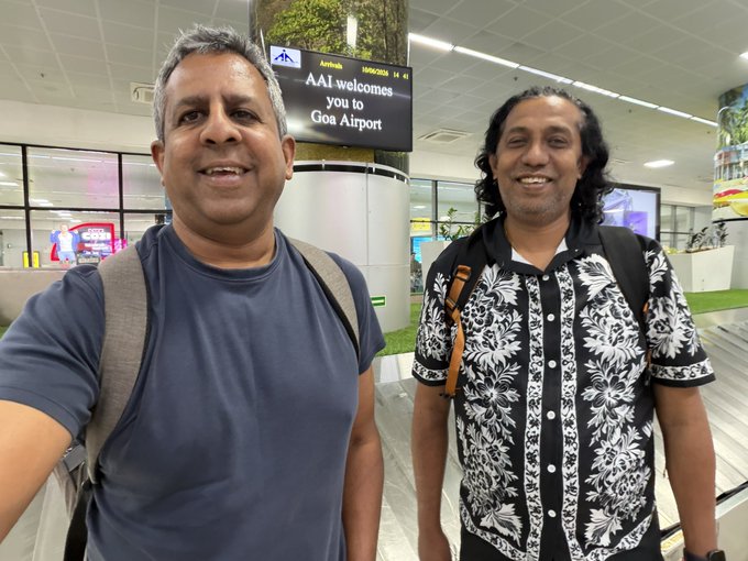 The image shows two men standing next to each other at an airport terminal. They are both smiling for a photo, with one of them wearing a backpack. The background features a sign that reads "AAI welcome Goa Airport", indicating they have arrived in Goa. A caption below the image suggests that they might be attending an Algorand India Dev Retreat or planning to go swimming at a pool.