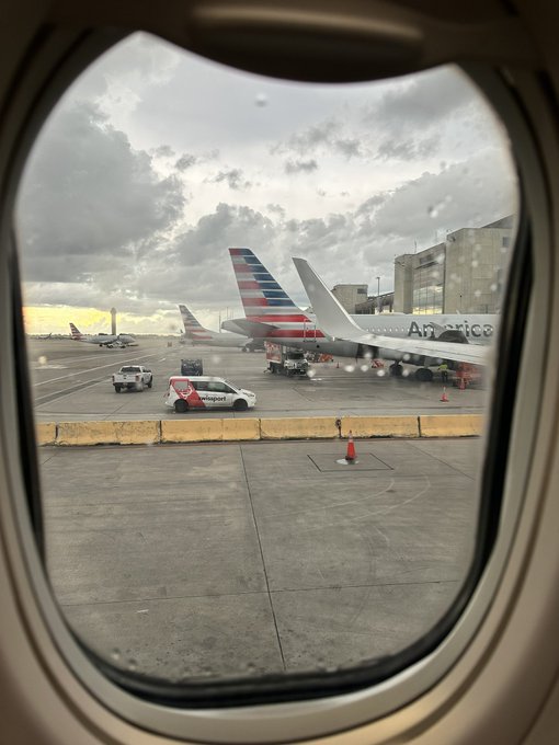 View through an airplane window showing American Airlines aircraft parked at an airport tarmac with ground service vehicles and traffic cones visible.