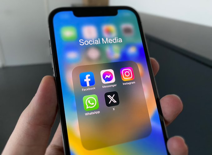 A close-up photograph of a hand holding a smartphone displaying a folder named 'Social Media' containing icons for Facebook, Messenger, Instagram, WhatsApp, and X.