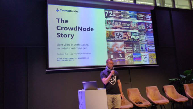 A man with a microphone presents on stage at a "Dash Conference" in Amsterdam (September 2024), standing beside a podium and a slide titled "The CrowdNode Story - Eight years of Dash Staking, and what must come next" by Andreas Rüdi. The screen also displays a collage of vintage-style promotional numbers like "50" and "43," fitting the event's crypto/Dash blockchain theme referenced in the post.