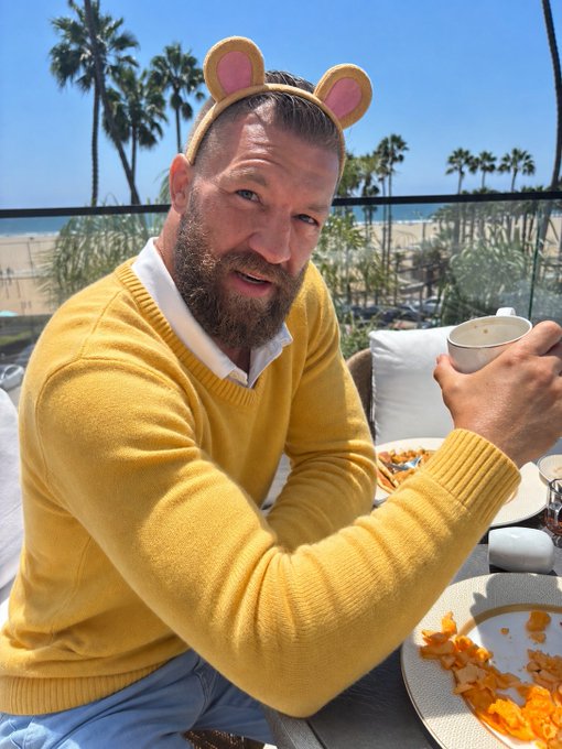 A man with a beard wearing a yellow sweater and a bear ear headband holds a teacup at an outdoor dining table with palm trees visible in the background.