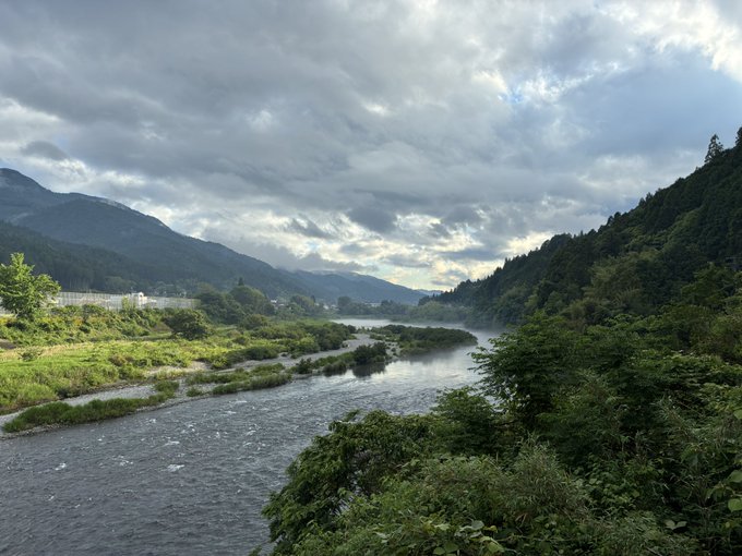 The image shows a river flowing through a lush green valley, with mountains visible in the background under an overcast sky. The water appears calm, reflecting the surrounding greenery. There are no people or other objects present in the scene, giving it a serene and peaceful atmosphere.
