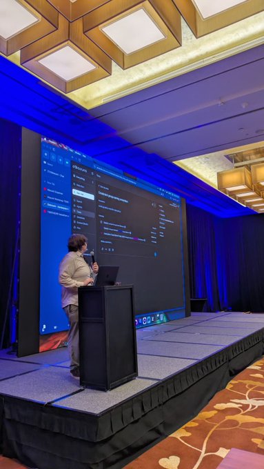 A presenter stands on a conference stage at the Token2049 hackathon, giving a live demonstration on a massive screen showing what appears to be an AI agent interface with the word "Masumi" visible at the top, while he speaks into a microphone next to a laptop on a podium. The scene captures the moment described in the post, with blue stage lighting and an empty auditorium setting emphasizing the live coding presentation.