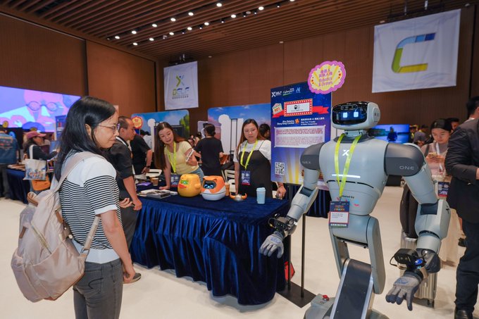 A woman observes the "ONE" humanoid robot at a Digital Entertainment Leadership Forum exhibition booth, which also features merchandise including a plush "Minibot" toy, matching the post's description of showcasing robots and the Floki Minibot at the event.