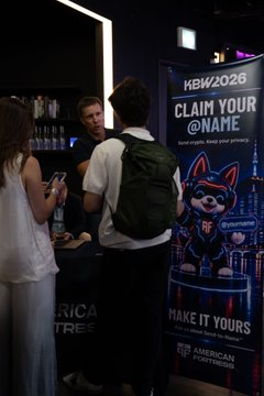 Two people stand near a registration booth beside a banner for 'KBW2026' featuring a blue dog mascot and the text 'CLAIM YOUR @NAME'.