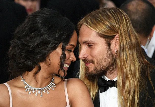 A woman with dark hair and a diamond necklace stands close to a man with long blonde hair and a beard, smiling at each other in formal attire at what appears to be a red carpet event.