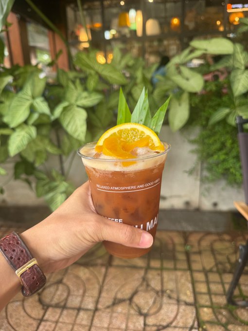 A hand wearing a watch holds a plastic cup of iced coffee garnished with an orange slice, with green plants in the background.